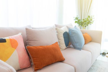 Modern oturma odasındaki beyaz kumaş kanepenin üzerinde turuncu ve mavi yastıklar ve tencerede yeşil ev bitkisi..