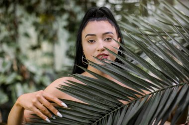 Doğal güzellik ve insan konsepti. Siyah at kuyruklu, gülümseyen genç Asyalı kadın, kameraya tropik bitkilerin arka planında poz veriyor, doğal palmiye yapraklarına dokunuyor.