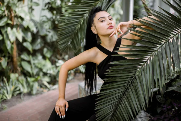 Retrato tropical de moda. Jovem modelo mulher asiática atraente em macacão preto na moda, com cabelo de rabo de cavalo, sentado perto de folhas de palma verde, enquanto posando em belas plantas tropicais de estufa — Fotografia de Stock