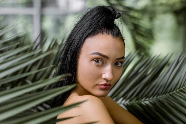 Close up retrato de beleza de jovem encantadora mulher asiática chinesa com estilo de cabelo de rabo de cavalo, posando para a câmera enquanto está perto de grandes folhas de palmeira exóticas em estufa ou jardim — Fotografia de Stock