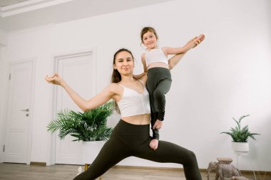 Yaşam gücü ve yoga yaşam tarzı konsepti. Zayıf, sportif bir anne ve aynı spor kıyafetleri giyen sevimli, neşeli bir kız, yoga stüdyosunda esneme egzersizleri yapıyor. Yoga pozisyonunda bir anne kızını kucağında tutuyor.