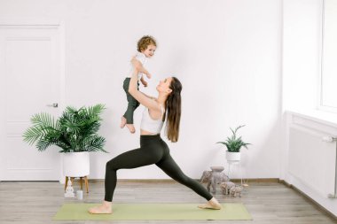 Spor giyim bölümünde anne ve küçük oğlu evde yoga yapıyorlar. Güzel, genç, beyaz bir anne oğluyla yoga yapıyor, anne yoga pozunda çocuğunu tutuyor.