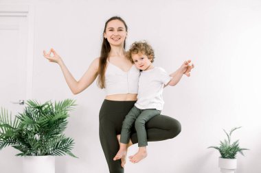 Gülümseyen anne ve oğlu yoga ağacı pozu ve mudra jesti yaparken, anne gülümseyen çocuğu kucağına alır. Genç anne ve oğlu hafif modern fitness stüdyosunda yoga yapıyor.
