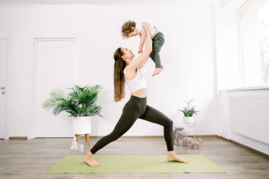 Şirin küçük oğlu ve güzel genç annesi sıcak yoga stüdyosunda ya da evde spor ve yoga egzersizleri yapıyor. Çekici sporcu kadın yeşil yoga minderinde duruyor ve küçük oğlunu büyütüyor.