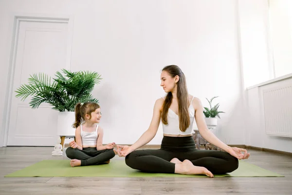 Pretty Caucasian mom with cute kid daughter doing yoga exercise at home or studio, sitting in lotus pose on the mat together. Mother teaching child to meditate and yoga