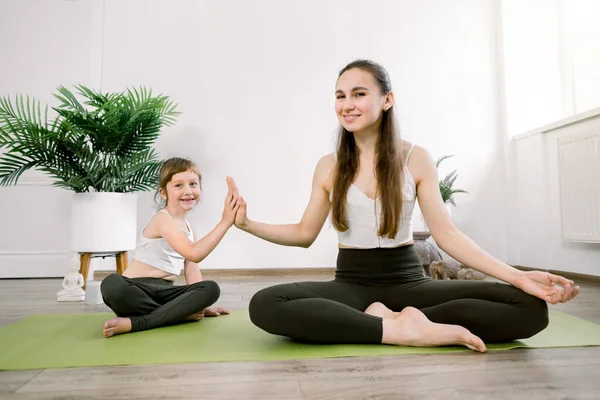 Mãe jovem e filha fofa, fazendo exercícios de esportes de ioga em casa, se divertir e passar um bom tempo juntos, sentados na posição de lótus e dando cinco altos. Conceito de um estilo de vida saudável — Fotografia de Stock