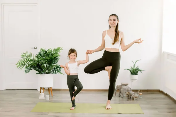 Concept of friendly family and yoga exercises. Pretty young mother and daughter doing yoga Tree pose. Woman and little girl are smiling while doing yoga together in fitness hall — Stock Photo, Image