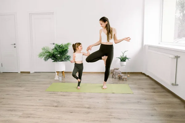 Front view of young mother having workout together with her little daughter, standing in tree yoga pose on green mat at cozy fitness hall. Sport, motherhood and active lifestyle concept. — Stock Photo, Image