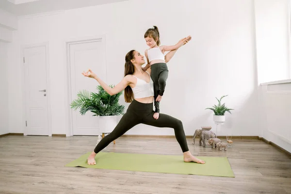 Attractive young Caucasian woman and her cute little daughter are smiling while doing yoga together at home. Pretty mom stands in Warrior One position and holds her little child girl — Stock Photo, Image