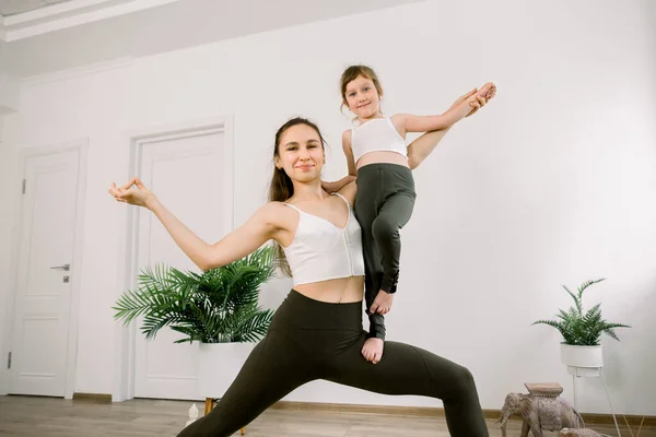 Vitality and yoga lifestyle concept. Slim sportive mom and cute cheerful joyful girl, wearing similar sportswear, doing stretching exercises in yoga studio indoors. Mom in yoga pose holds her daughter — Stock Photo, Image