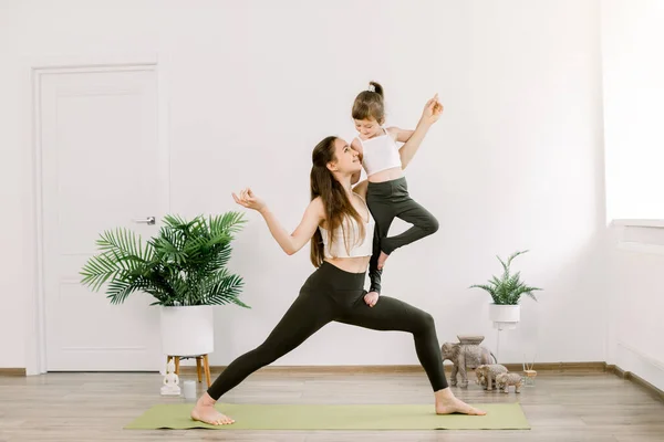 Young mom and daughter doing yoga gymnastics on green mat in light cozy room at home. Woman standing on yoga position holds her daughter in tree pose, looking each other