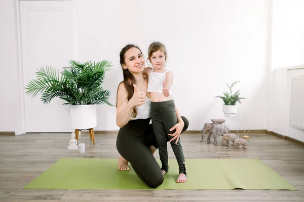 Esporte familiar e conceito de ioga. Promoção do estilo de vida saudável. Mãe e pequena filha sorrindo juntos para a câmera e mostrando os polegares para cima, enquanto em pé no estúdio de ioga luz aconchegante — Fotografia de Stock