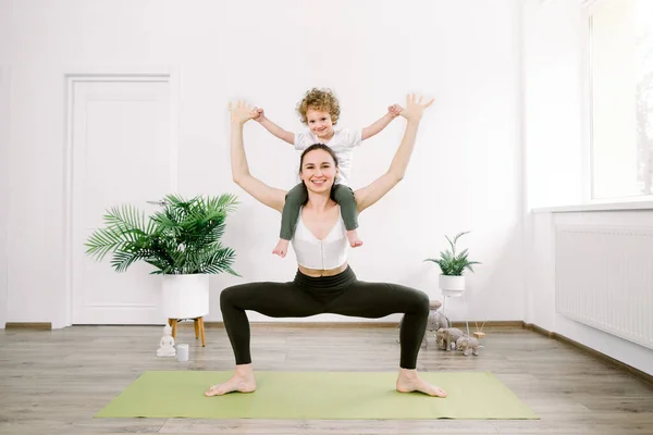 Beautiful young Caucasian woman and her little son are smiling while doing yoga together at home. Woman in yoga position with her son sitting on her shoulders — Stock Photo, Image