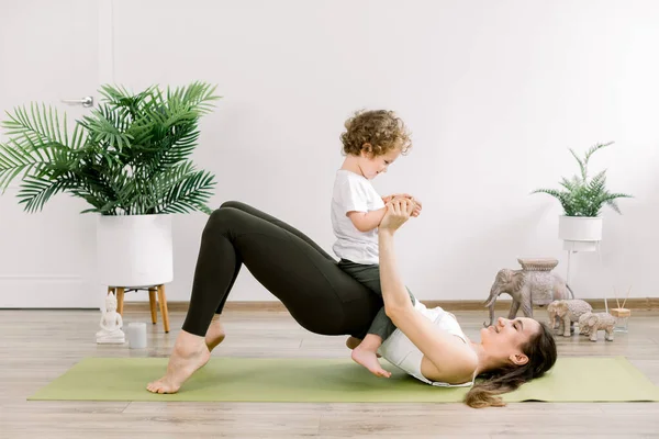 Young happy sporty woman mother working out, doing butt bridge pilates yoga exercise, little kid son on her belly. Healthy lifestyle, fitness, postnatal yoga, maternity concept — Fotografia de Stock