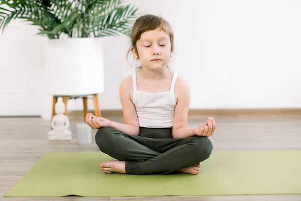 Маленькая хорошенькая девочка расслабляется с закрытыми глазами в позе лотоса йоги, сидя дома на зеленом коврике. Симпатичная девушка сидит с закрытыми глазами в позе лотоса и жестом мудры