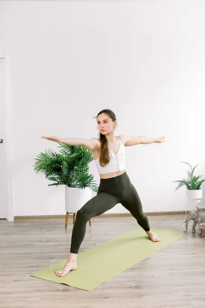 Vista de ángulo lateral de la mujer deportiva practicando yoga, de pie en Guerrero dos ejercicios, Virabhadrasana II posan en la esterilla verde, haciendo ejercicio en el estudio de yoga ligero. Longitud total. Concepto de yoga y salud —  Fotos de Stock