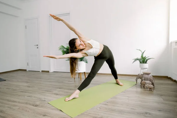 Horizontal indoor shot of pretty young fitness yoga couch, standing in Floating Triangle yoga pose on green mat, enjoying her sporty time in the morning — Stock Photo, Image