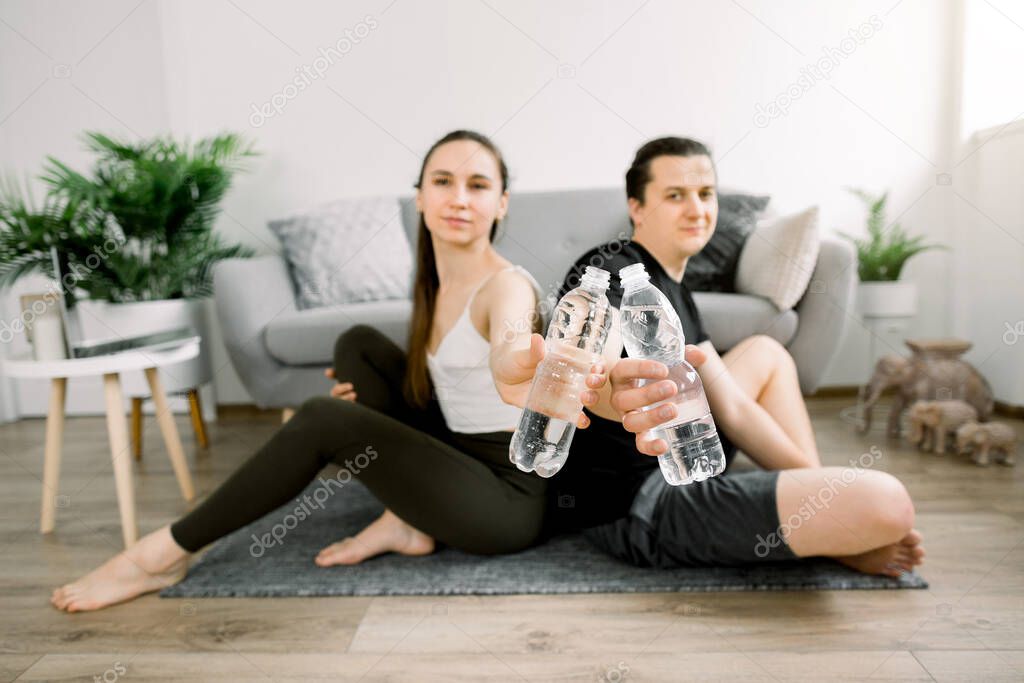 Sporty young man and woman, sitting on the floor back to back, resting after home workout, showing water in plastic bottles to camera. Water balance concept. Focus on the bottles