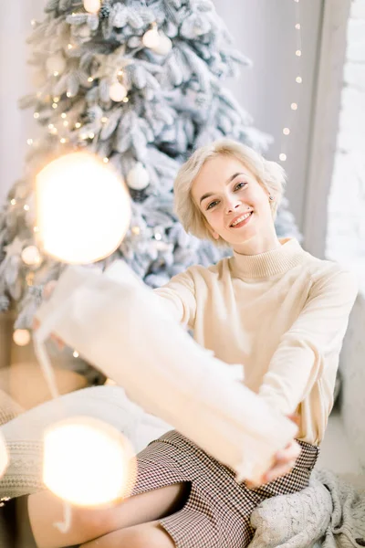 Weihnachten, Weihnachten, Winter, Geschenke und Glückskonzept. Lächelnde blonde Frau in gemütlichen warmen trendigen Kleidern, sitzt am geschmückten Raum mit Lichtern und Weihnachtsbaum mit Geschenkbox — Stockfoto