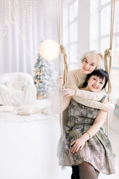 Beautiful Caucasian girl in dress, sits on the swing at cozy light decorated studio with Christmas and New year decorations, fir tree and big window, while her pretty female friend hugs her — Stock Photo, Image