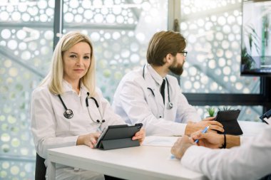 Başarılı gülümseyen, beyaz, sarışın, orta yaşlı bayan doktor, tablet bilgisayar kullanıyor, iş arkadaşlarıyla ışık odasında otururken gülümsüyor ve çevrimiçi video konferansı yapıyor.
