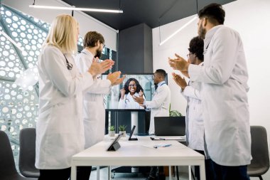 Uluslararası profesyonel hekimler modern klinikteki toplantı odasında el çırpıyor, heyecanlı Afrikalı kadın baş doktorlarıyla görüntülü sohbet ediyor, başarıyı kutluyor, yumruklarını sıkıyor.