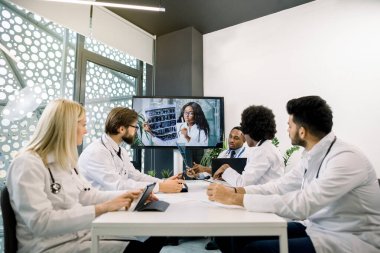 Bir grup çok ırklı doktor masada oturup bilgisayar ekranındaki genç Afrikalı bayan meslektaşlarını dinliyor ve çevrimiçi konferans sırasında bilgisayarlı tomografi sonuçlarını tartışıyorlar.