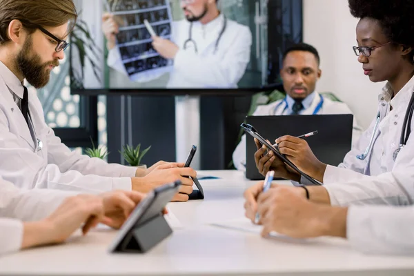 Rajattu kuva joukkue monirotuinen lääkärit käyttävät videonäyttö projektori konferenssin aikana sisätiloissa, heidän kollegansa keskustella CT. Afrikkalainen nainen ja valkoihoinen mies tekee muistiinpanoja — kuvapankkivalokuva