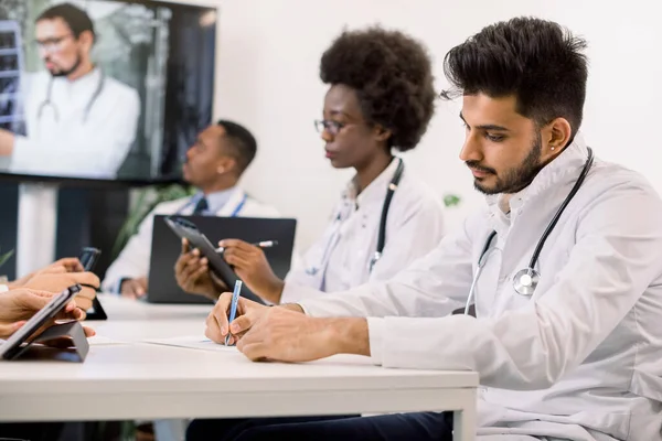 Gruppo di medici multietnici maschili e femminili che utilizzano appunti e laptop, prendendo appunti durante la videoconferenza online con lo scienziato caucasico. Concentrati sugli appunti dei medici indiani — Foto Stock