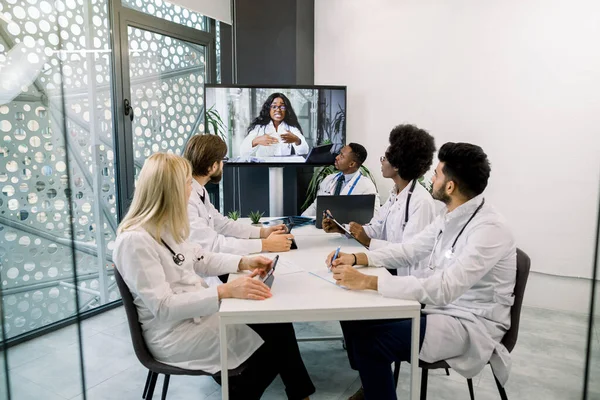 Squadra di cinque medici multietnici che si riuniscono in videoconferenza in ospedale con la loro giovane dottoressa africana. Squadra di assistenza sanitaria su webinar incontro online. Concetto di telemedicina. — Foto Stock