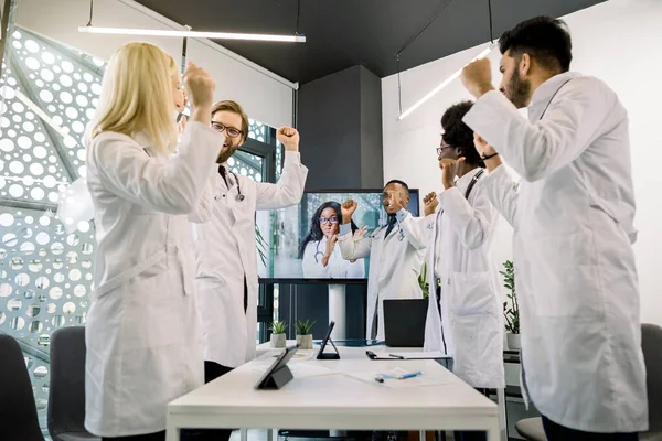 Medicine, conference, victory, successful trials. Five excited cheerful multiethnic doctors celebrating success, clenching fists and shouting, having video chat with their African female colleague