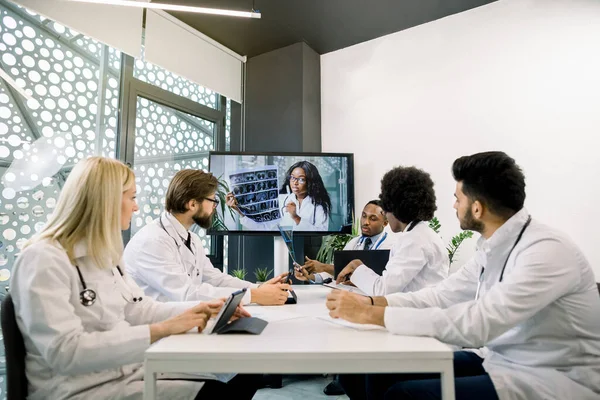 Sekelompok dokter multietnis duduk di meja dan mendengarkan rekan wanita muda Afrika mereka di monitor komputer, membahas hasil tomografi yang dihitung selama konferensi online — Stok Foto