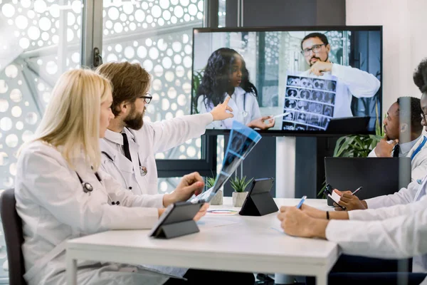 Team of multiethnic doctors looking at the screen in video conference meeting at hospital, chatting with two confident African and Caucasian doctors, holding CT and discussing the treatment
