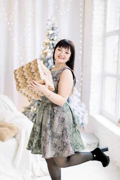 Retrato de sorrindo jovem mulher morena caucasiana elegante em vestido de luxo olhando para a câmera enquanto segurava a caixa de presente de Natal perto da árvore de Natal no quarto decorado aconchegante branco — Fotografia de Stock