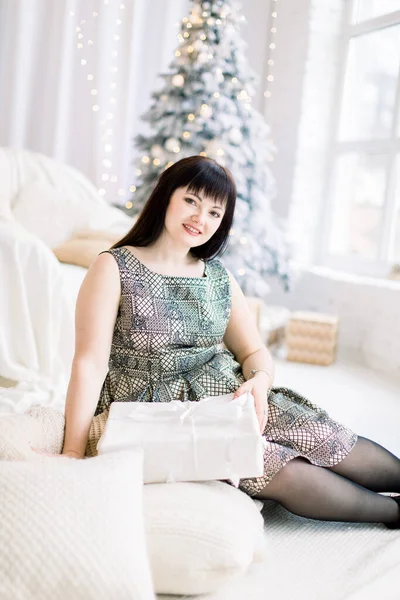 Menina bonita no vestido está sentado no chão entre almofadas brancas e segurando caixa de presente. Acolhedor quarto decorado com árvore de Natal e sofá no fundo — Fotografia de Stock