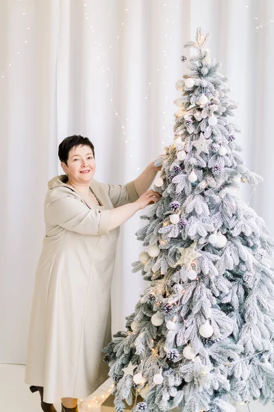 Winter holidays, Christmas and New Year concept. Happy middle aged brunette woman in elegant dress preparing for Christmas, decorating fir tree and posing to camera at cozy room at home