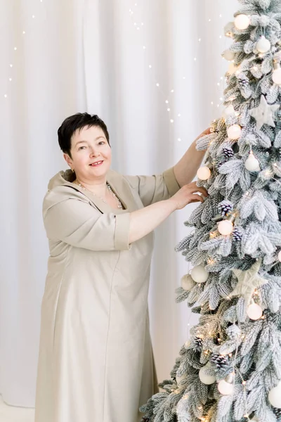 Close up retrato de mulher branca madura atraente, vestindo elegante vestido bege, decorando árvore de Natal por si mesma em casa. Conceito de Ano Novo e Natal — Fotografia de Stock