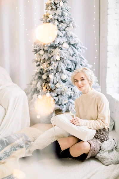 Bella giovane donna bionda in pullover e gonna a scacchi, si siede su plaid lavorato a maglia con cuscini sul pavimento, in bella stanza decorata interna con albero di Natale sullo sfondo e luci — Foto Stock