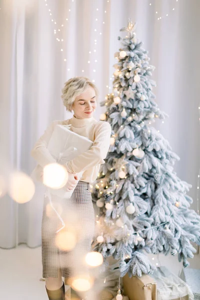 Jovem mulher loira sonhadora atraente, de pé na frente da árvore de Natal decorada e segurando caixa de presente de Natal. Jovem feliz bonita segurando um presente de Natal — Fotografia de Stock