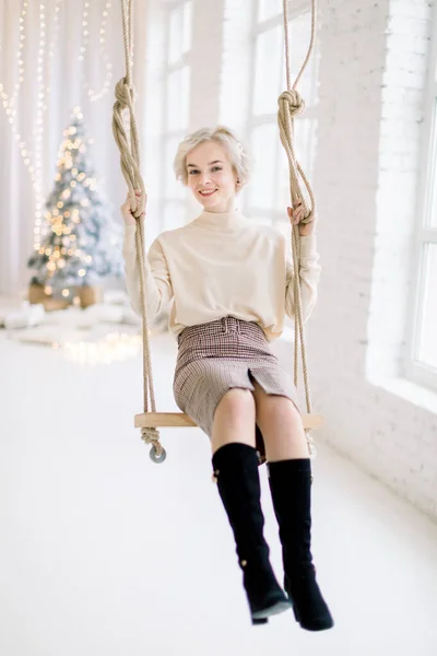 Menina loira encantadora jovem em roupas quentes da moda, balançando no balanço de madeira em belo quarto decorado luz com árvore de Natal e grandes janelas no fundo — Fotografia de Stock