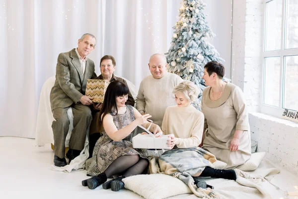 Cheerful big laughing European family with grandparents opening Christmas gifts, while sitting at cozy light decorated room with big windows and Christmas tree