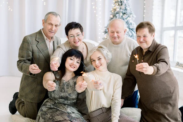 Joyeux sourire famille de trois générations, grands-parents, parents et deux sœurs, tenant des étincelles, assis devant l'arbre de Noël. Concept multi générations, Noël et Nouvel An. — Photo