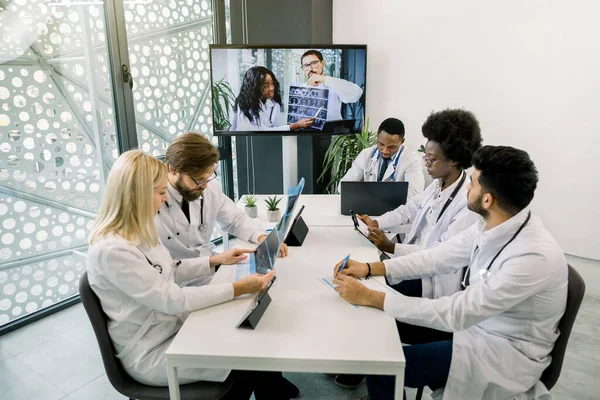 Tim yang terdiri dari lima orang dokter multietnis yang berbeda-beda mengadakan pertemuan konferensi video di rumah sakit, mengobrol dengan dua rekan muda Afrika dan Kaukasia mereka, menganalisis CT, cara pengobatan dan diagnosis — Stok Foto