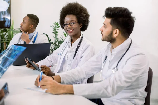 Beskuren bild av team av multietniska läkare, tittar läkare tal på digital skärm på videokonferens i ljusrummet. Fokusera på ganska leende afrikanska kvinnliga läkare, att göra anteckningar — Stockfoto