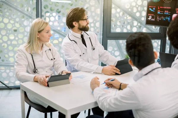 Tıp personeli toplantısı veya klinikteki konferans sırasında masanın etrafında oturan çok ırklı genç doktorların yan açılı görüşleri ve dijital ekrana çizelgeler ve şemalarla bakmaları — Stok fotoğraf