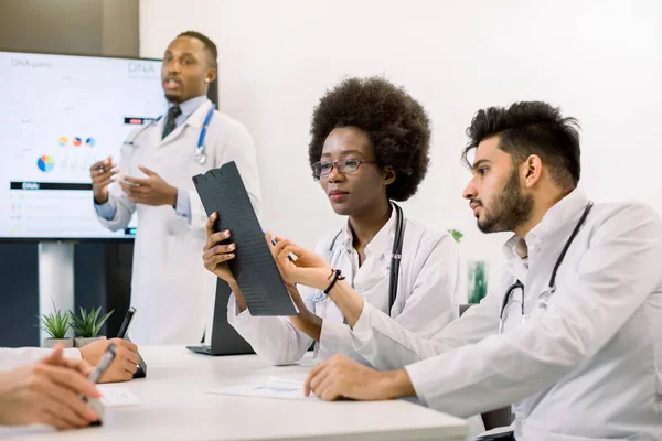 İki genç doktorun portresi, Afrikalı kadın ve Hintli adam, masada oturup tıbbi durumu tartışıyorlar. Yakışıklı Afrikalı erkek doktor arka planda konuşma yapacak. — Stok fotoğraf