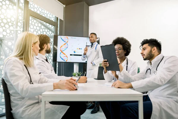 Cinq médecins multiethniques discutent pendant la réunion dans la salle de conférence, en utilisant des tablettes et des schémas. Beau médecin africain homme orateur, montrant strusture biochimique — Photo