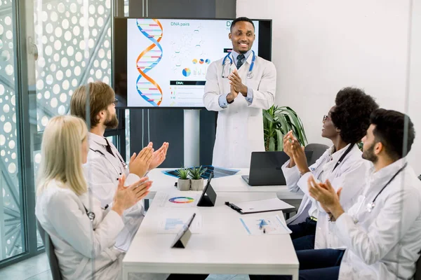Çok ırklı doktorlar, dijital ekranın yanında duran ve DNA yapısı ve yeni ilaçlar hakkında başarılı bir konuşma yapan neşeli ve yakışıklı Afrikalı meslektaşlarını alkışlıyor. — Stok fotoğraf
