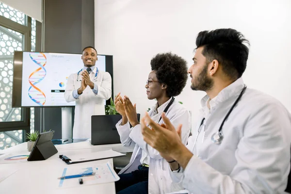 Giovane medico maschio africano che fa un discorso di successo per la squadra medica, mentre in piedi di fronte a grande schermo digitale con grafici e struttura del DNA. Giovani medici indiani e africani applaudono — Foto Stock