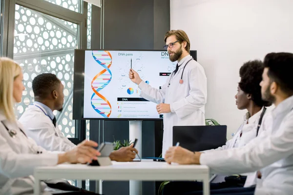 Beau jeune homme caucasien médecin scientifique portant un manteau blanc debout près de l'écran numérique et donnant une présentation sur les molécules biochimiques, de nouveaux médicaments pour le groupe multiethnique de ses collègues — Photo